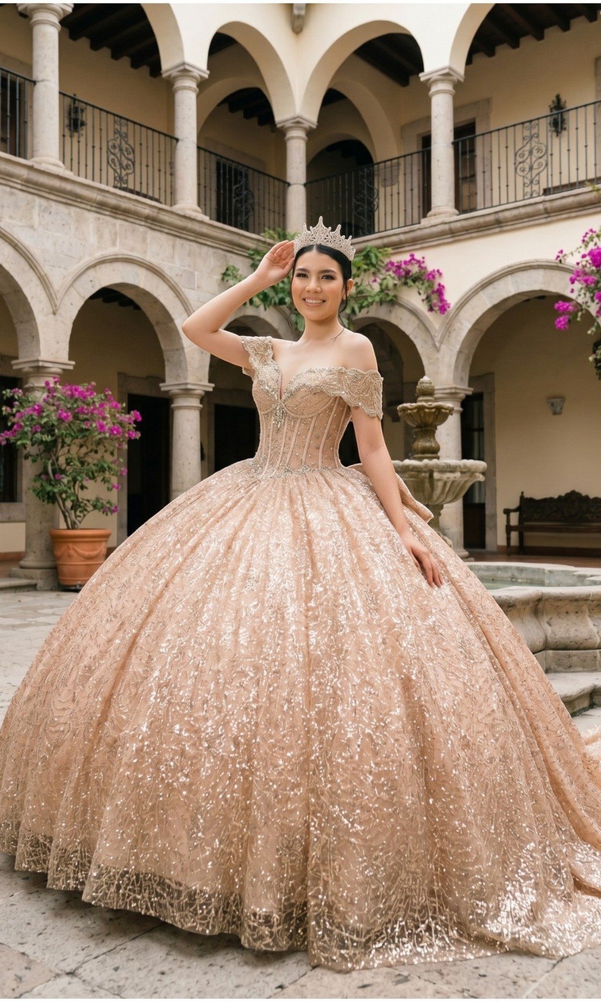 A young woman in the Quinceanera Dress 8600JQ By Cinderella Couture, an ornate, sparkling rosegold ball gown featuring a corset bodice and off-the-shoulder neckline, stands smiling at the camera in a courtyard with stone arches and flowering plants.